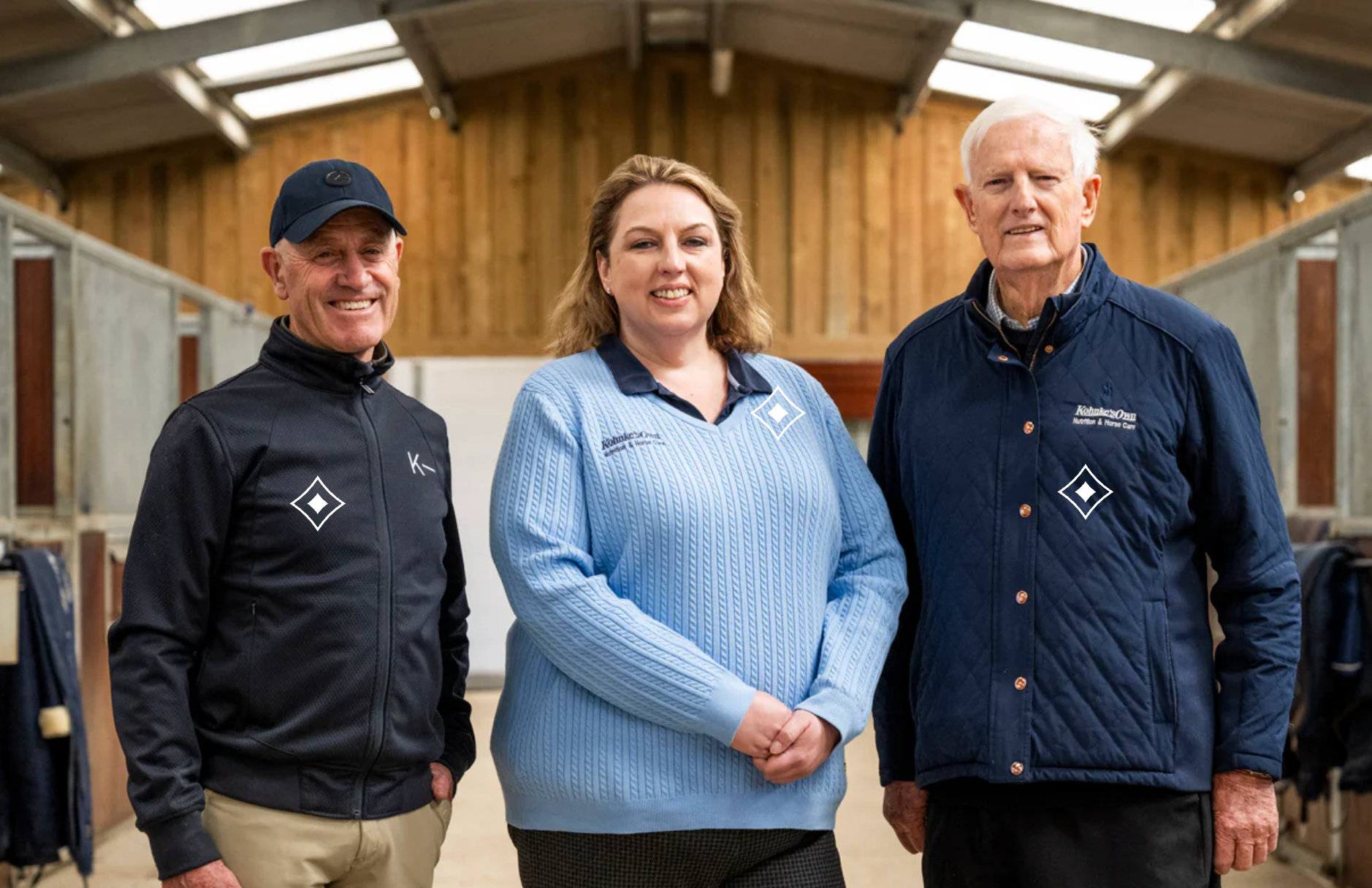 Three people standing in a stable. A man and a woman flank an older man. They wear blue branded jackets and sweaters.