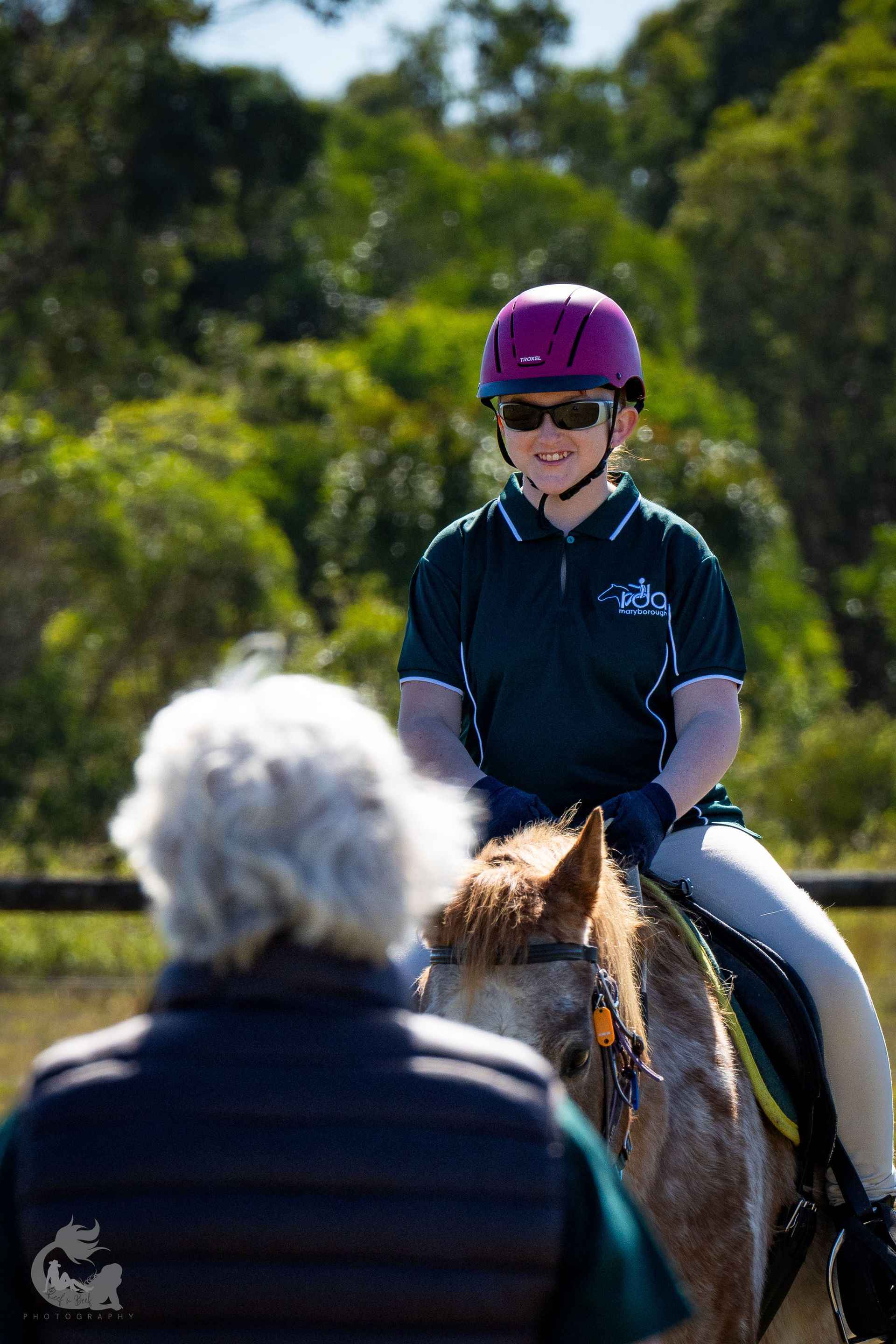 Person riding a horse, wearing helmet, being observed by another. Outdoors, sunny.