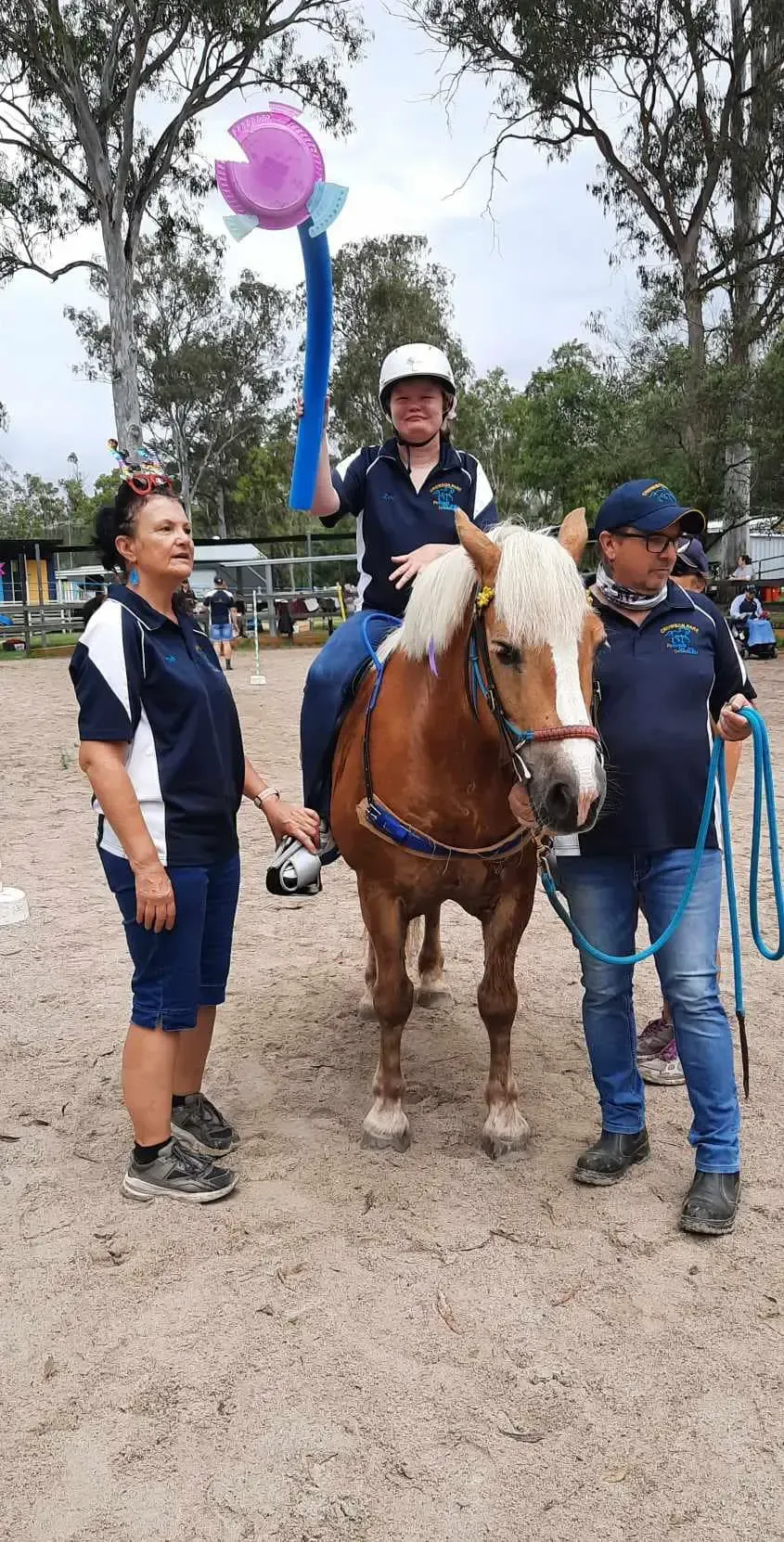Person on a brown horse, holding a trophy, flanked by two people, at an outdoor event.