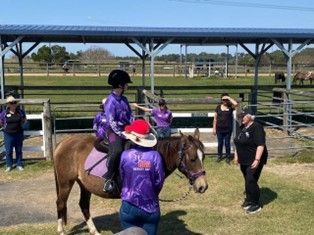 Child on a pony, wearing purple, assisted by people. Horse riding lesson outdoors, under a shelter.