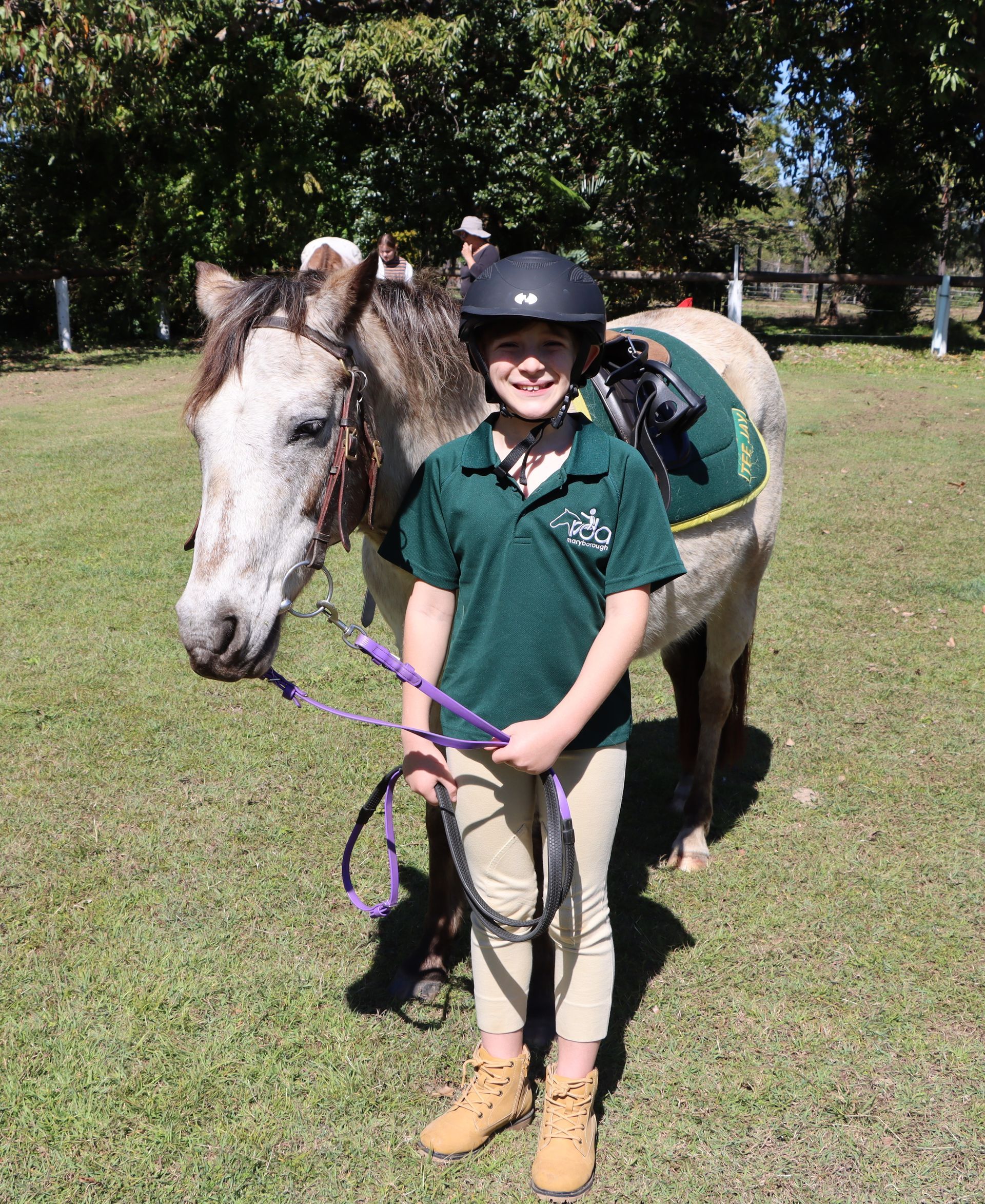 Young person in riding gear smiling next to a horse on grass.