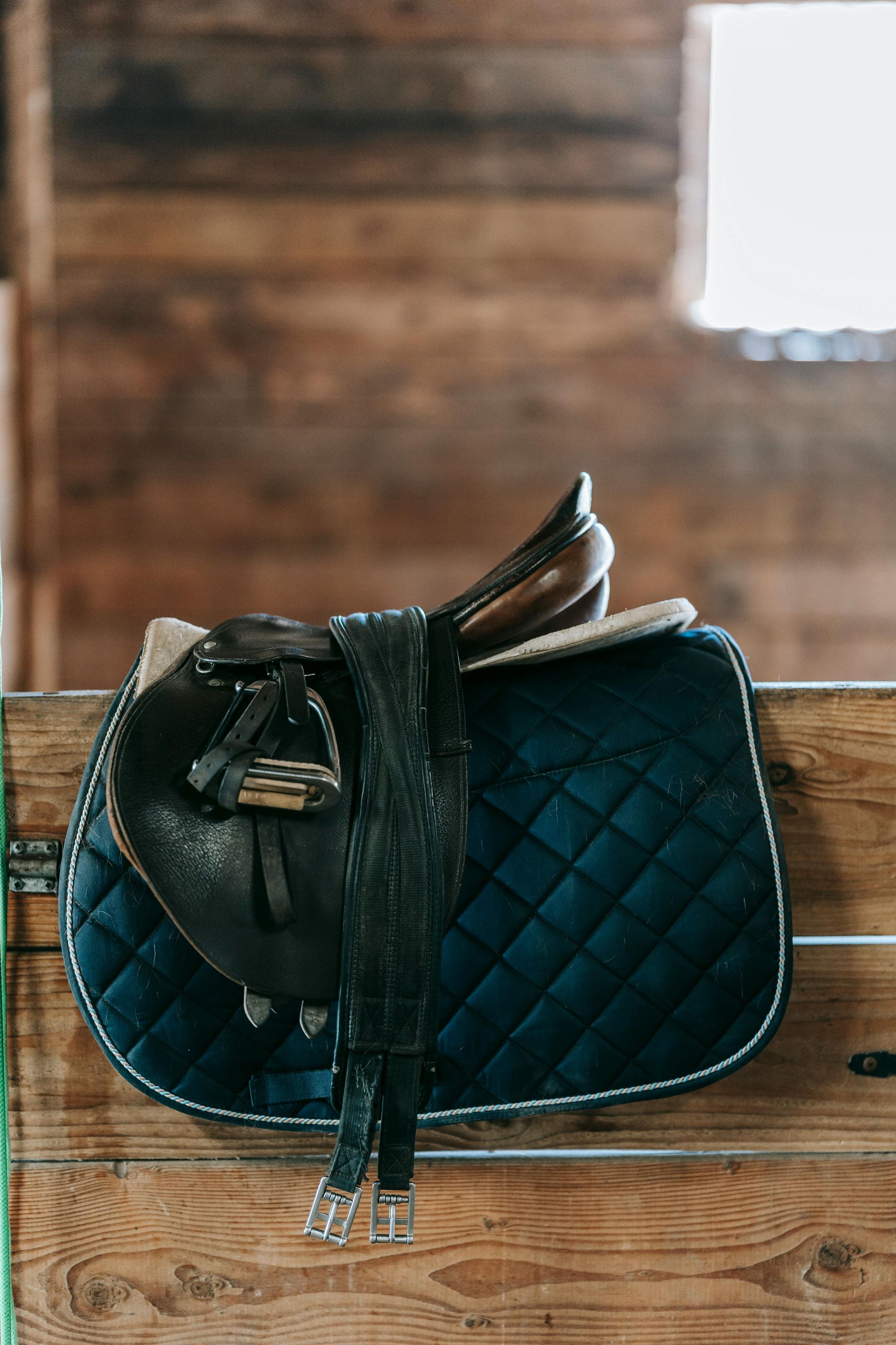 Leather horse saddle on navy quilted pad, wooden stall background.