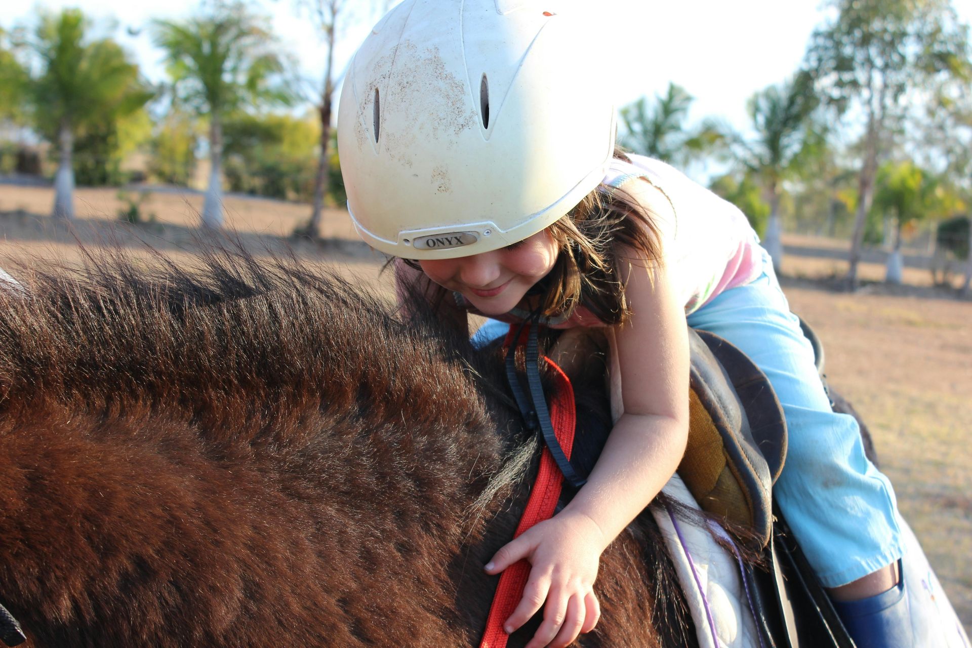 Girl in white helmet on a brown horse, smiling, petting the horse's mane outdoors on a sunny day.