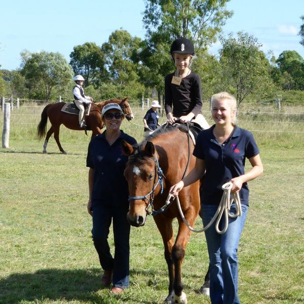 Two women lead a child riding a brown horse in a grassy field. Other children ride horses in the background.