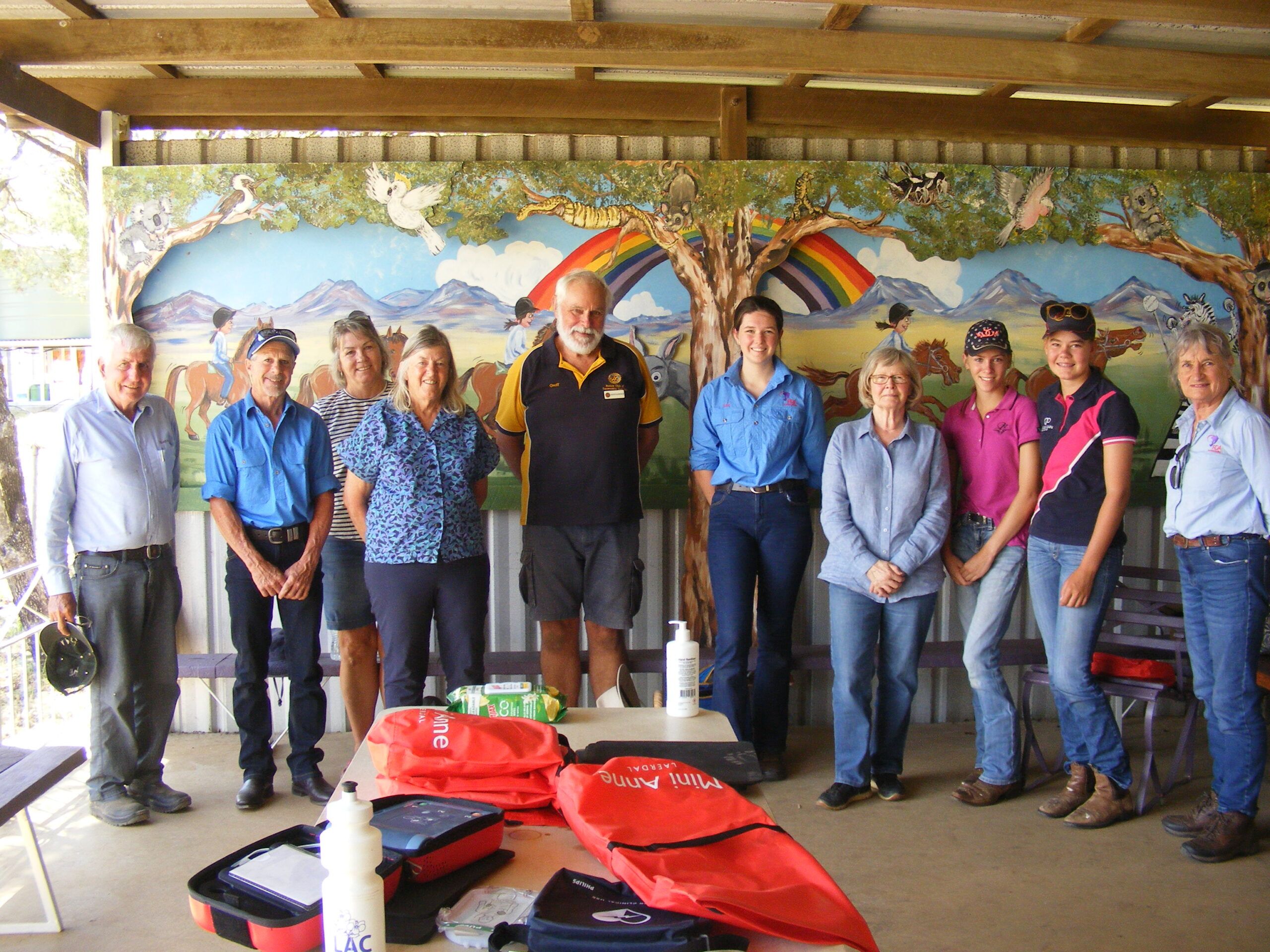 Group of people posing near a table with first aid kits; mural of a landscape with a rainbow in the background.