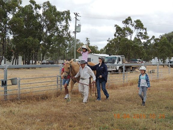 People riding a horse at an outdoor event. Two adults lead the horse while a child sits on its back. Another child walks nearby.