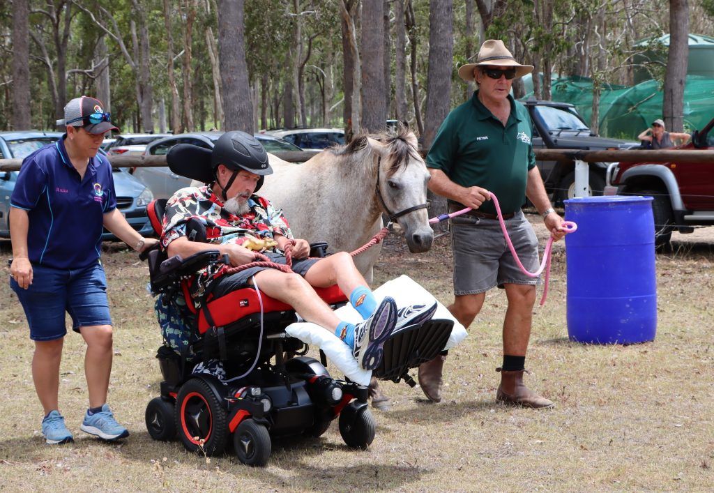 Man in wheelchair with horse, guided by two people in outdoor setting.