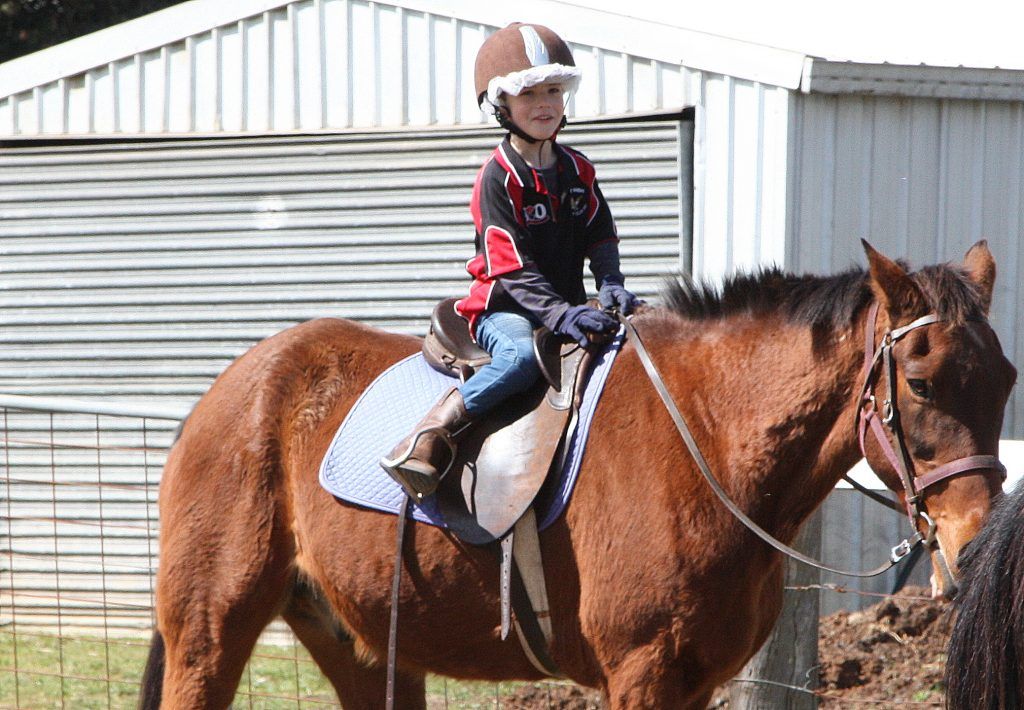 Child on a brown horse, wearing helmet and boots, in a stable area.