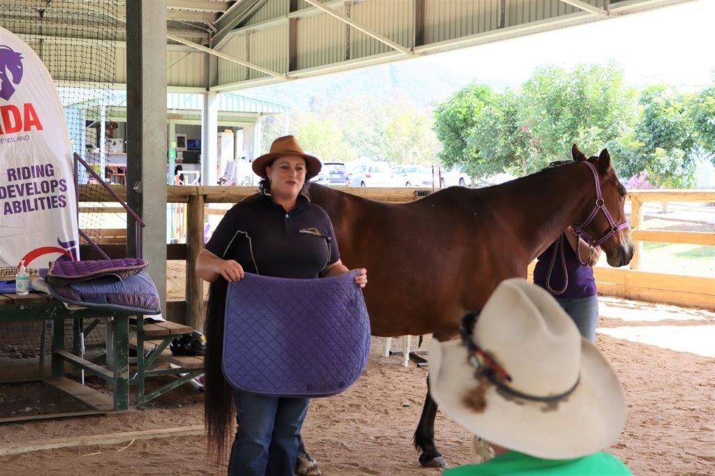 Woman in a cowboy hat holding a blue saddle pad, showing it to a horse and a seated audience in a stable.