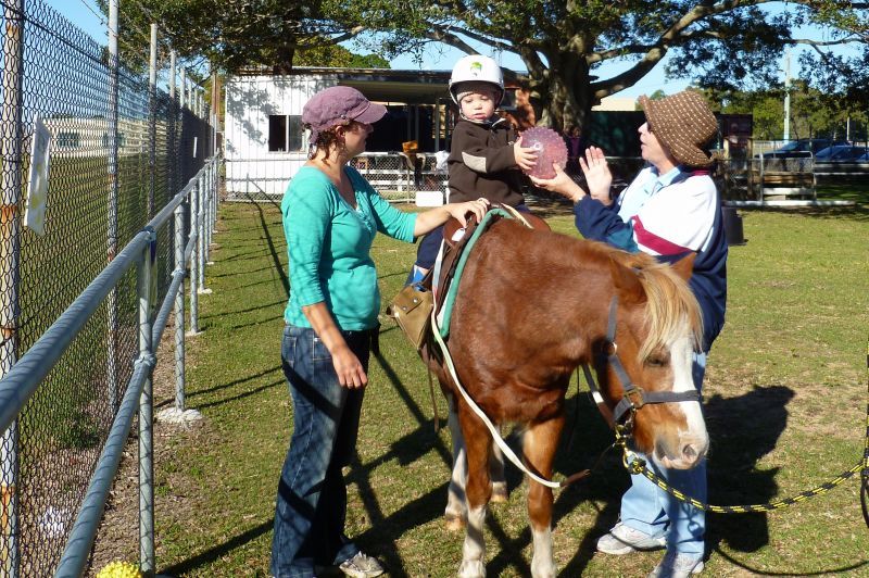 A child rides a pony, guided by two adults on a grassy area, fenced in.
