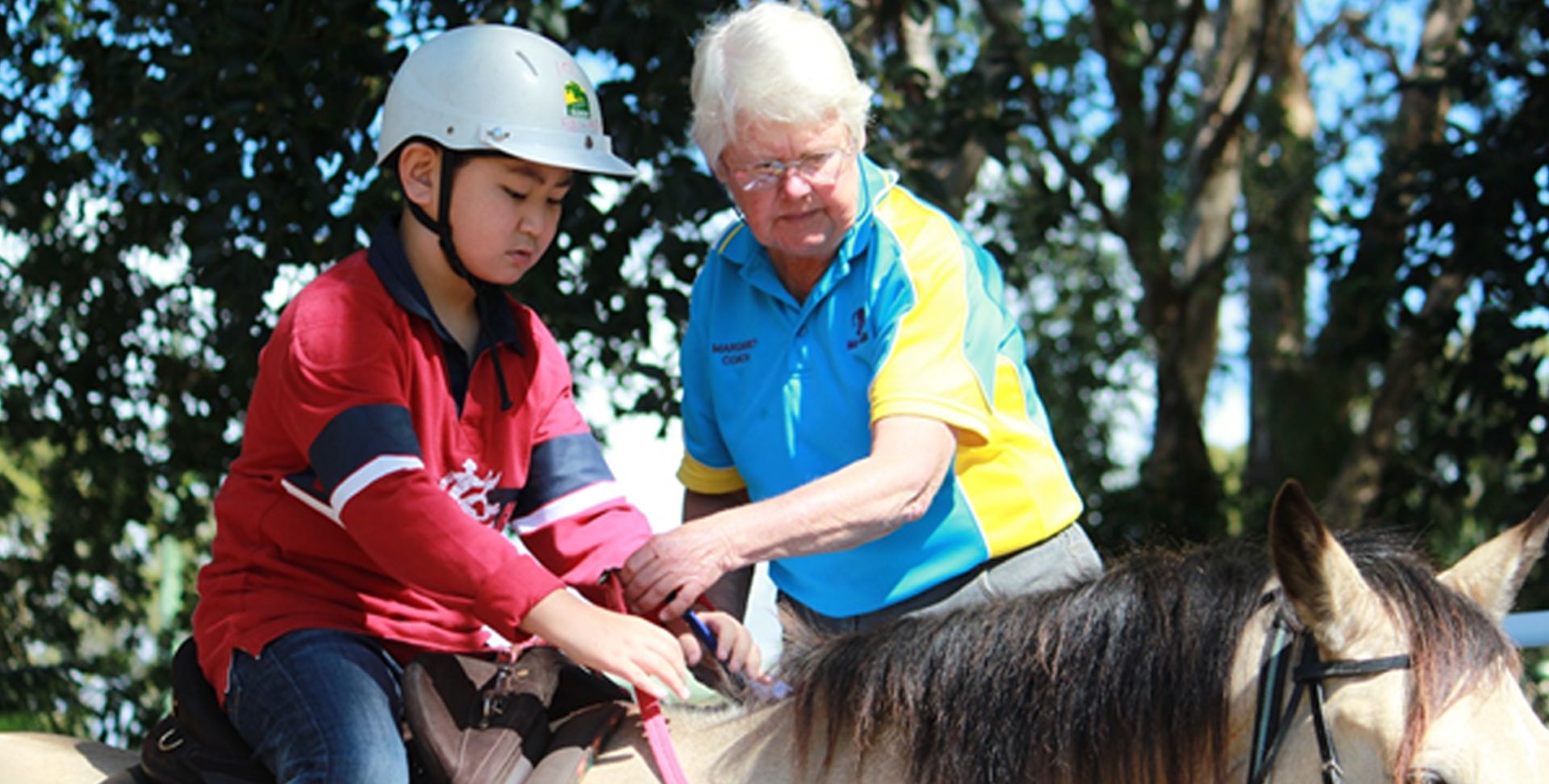 A person in a helmet adjusts the saddle on a horse with the help of an instructor.