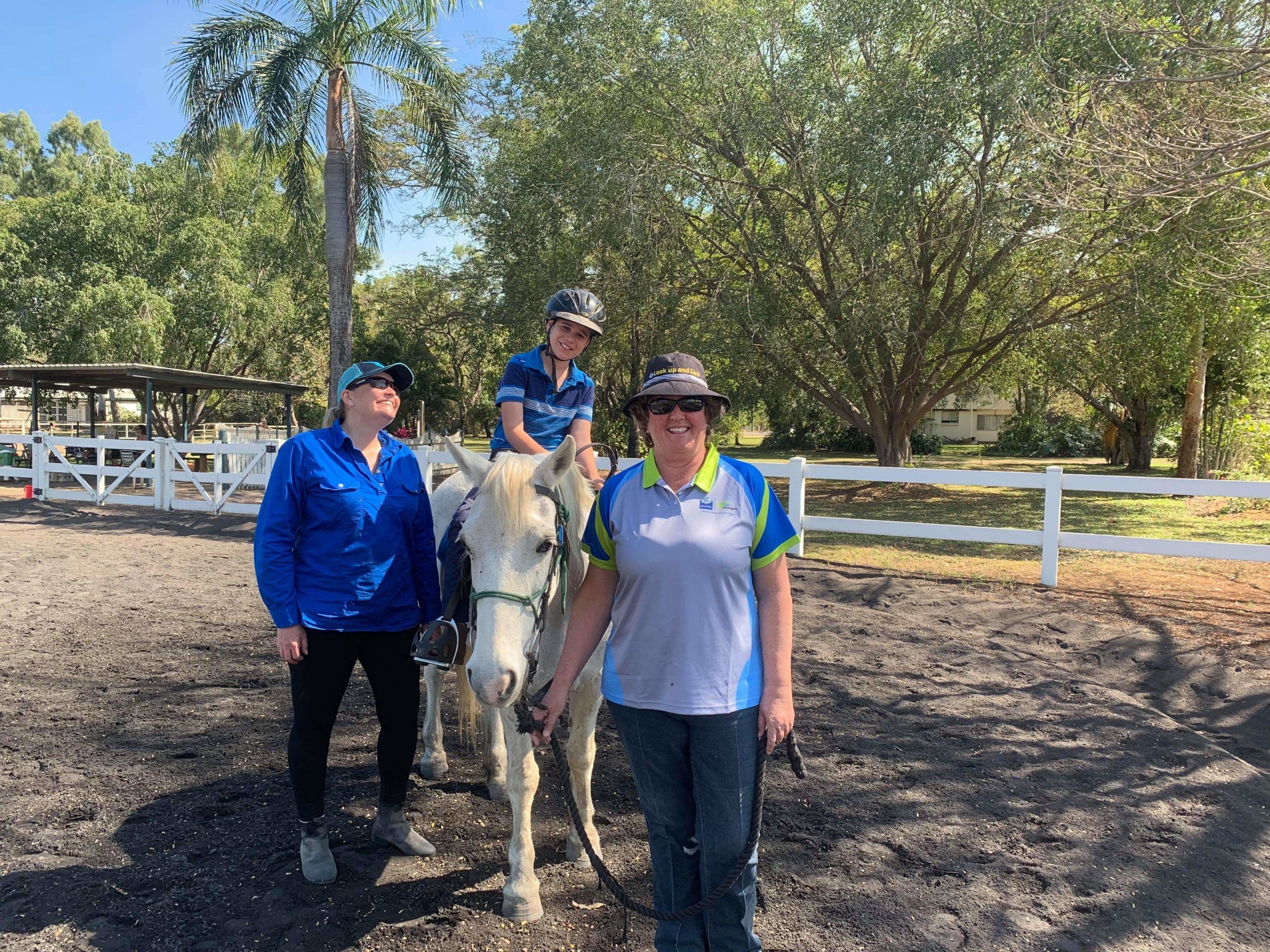 Person on horseback with two assistants in an outdoor riding area with trees and white fencing.
