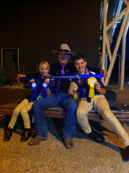 Man in cowboy hat with two kids, holding blue and yellow ribbons, seated on a bench at night.