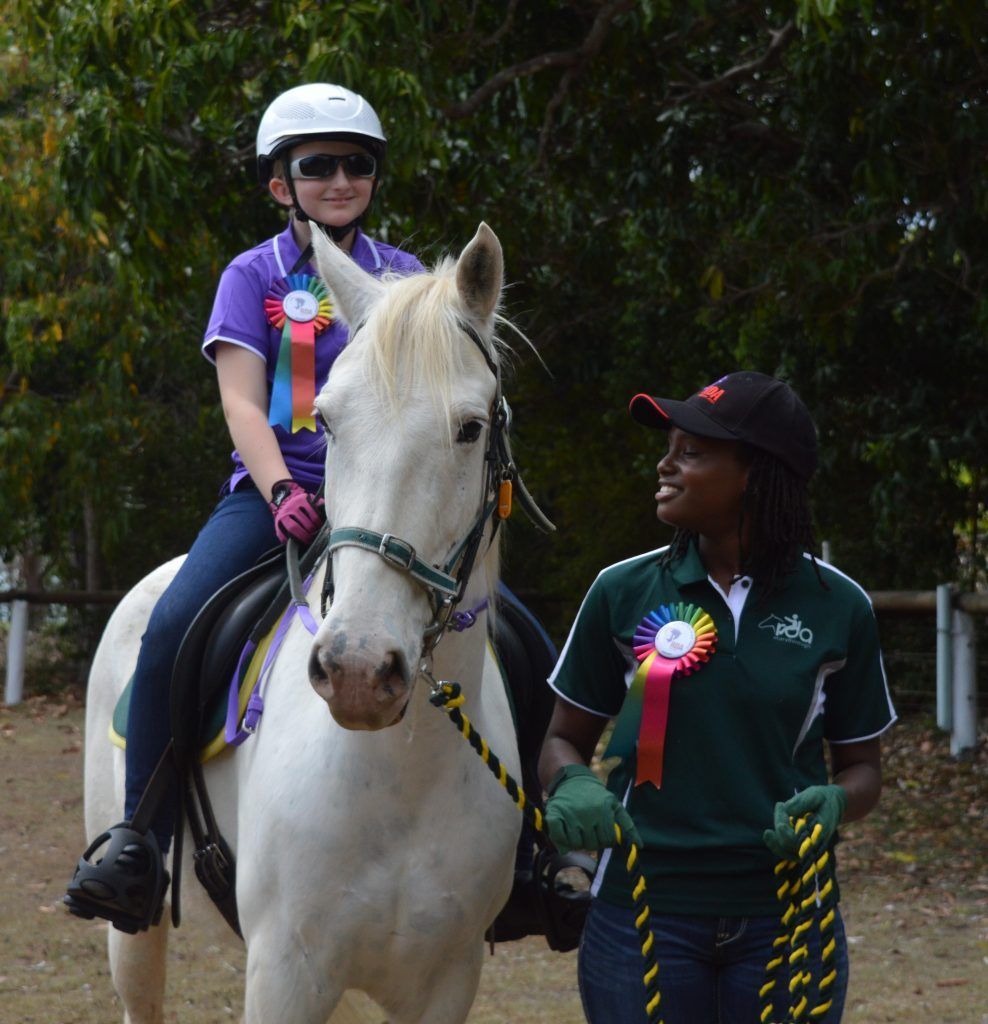 Girl on white horse, guided by smiling woman; outdoors, green shirt, riding helmets.