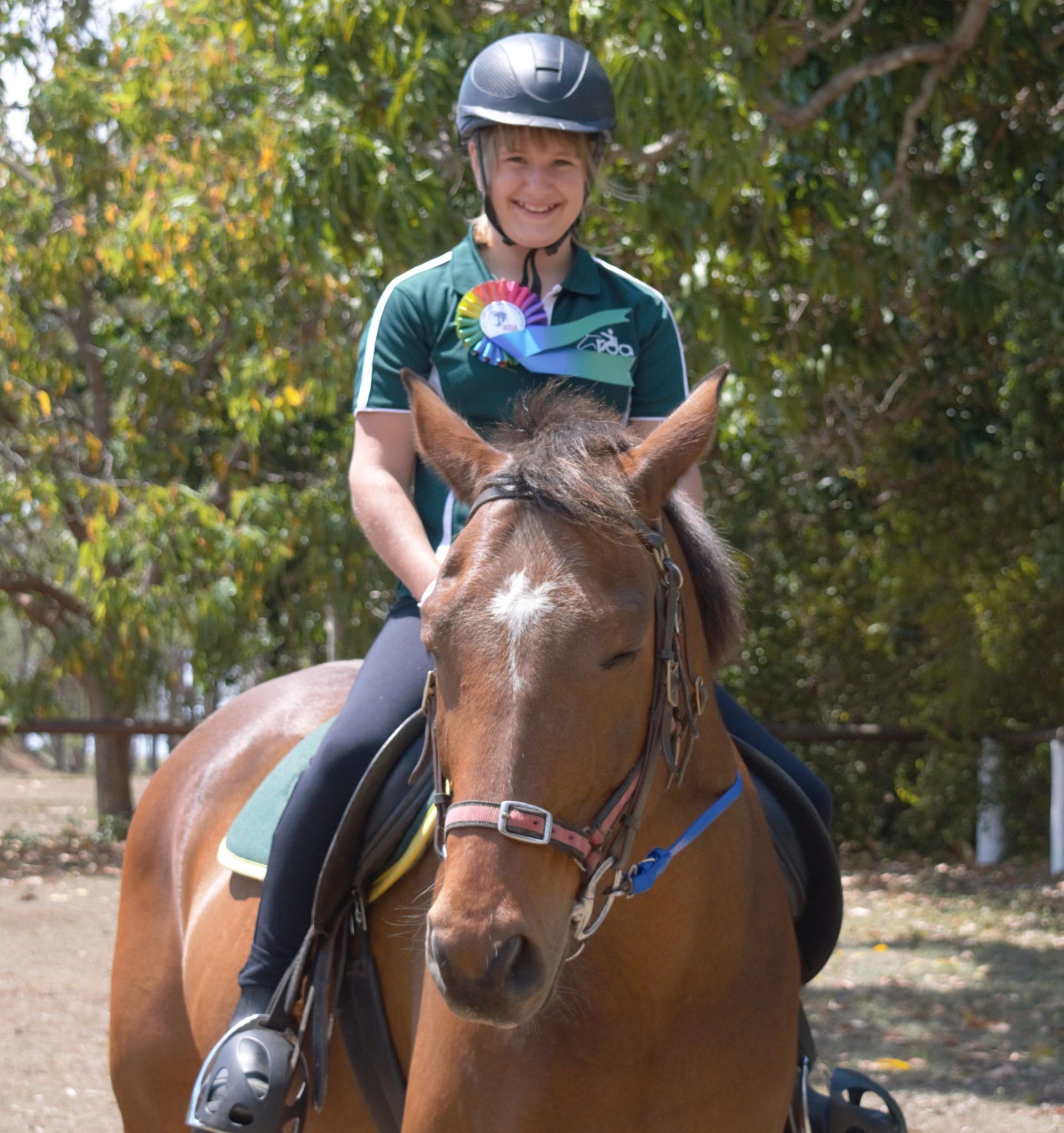 Woman riding brown horse in outdoor setting. Rider wearing helmet, green shirt, and blue ribbons.