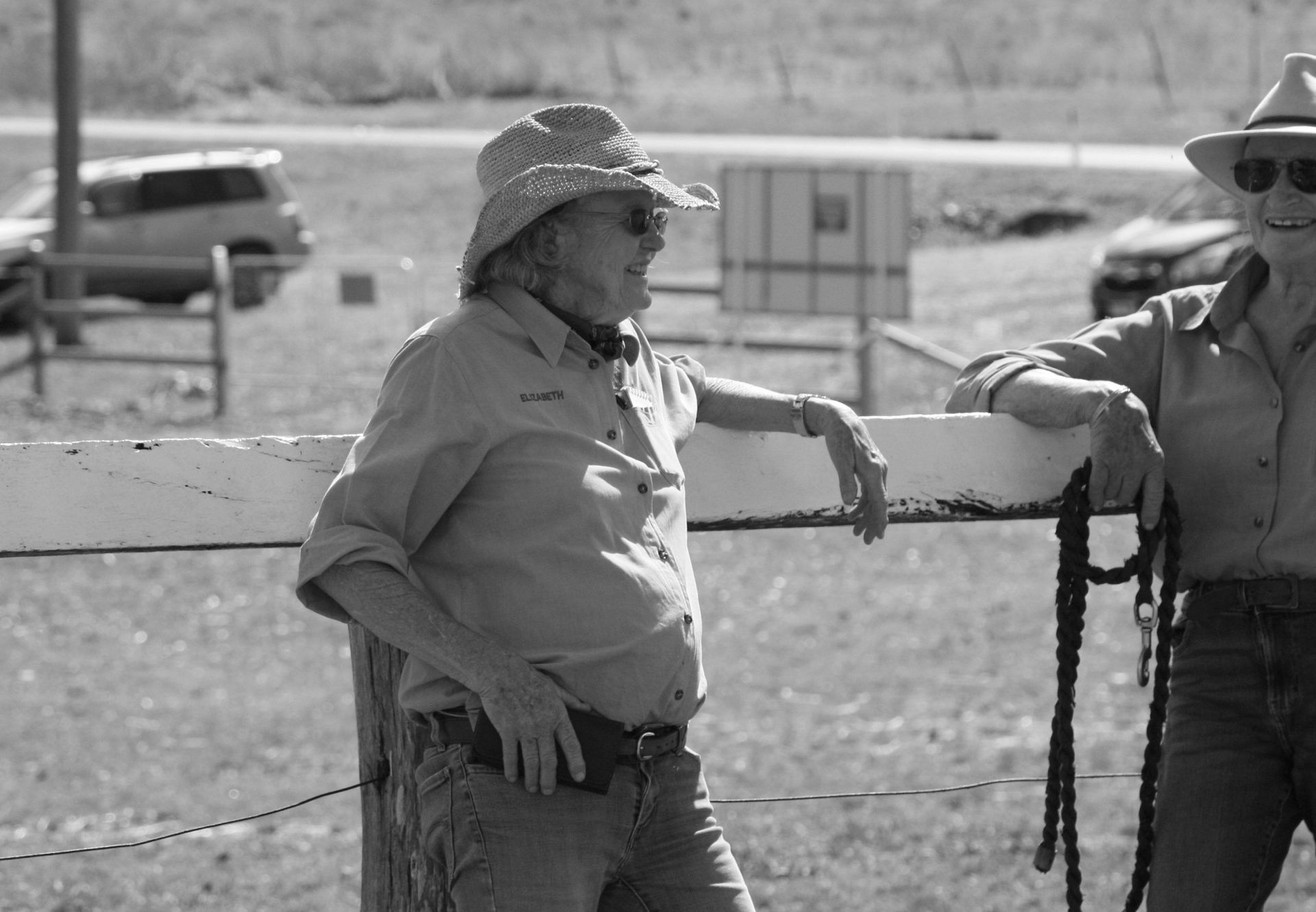 Two people wearing hats lean on a fence outdoors. One has a straw hat, the other sunglasses.