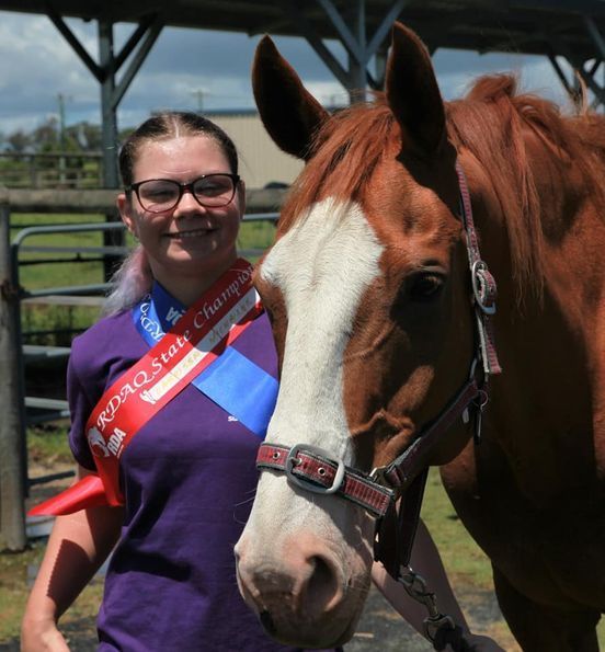 Teen with glasses and horse, both wearing prize ribbons. Outdoors, sunny.