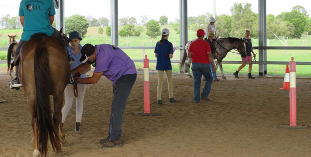 People with horses in a riding arena. A person adjusts a horse's tack. Others watch and ride.