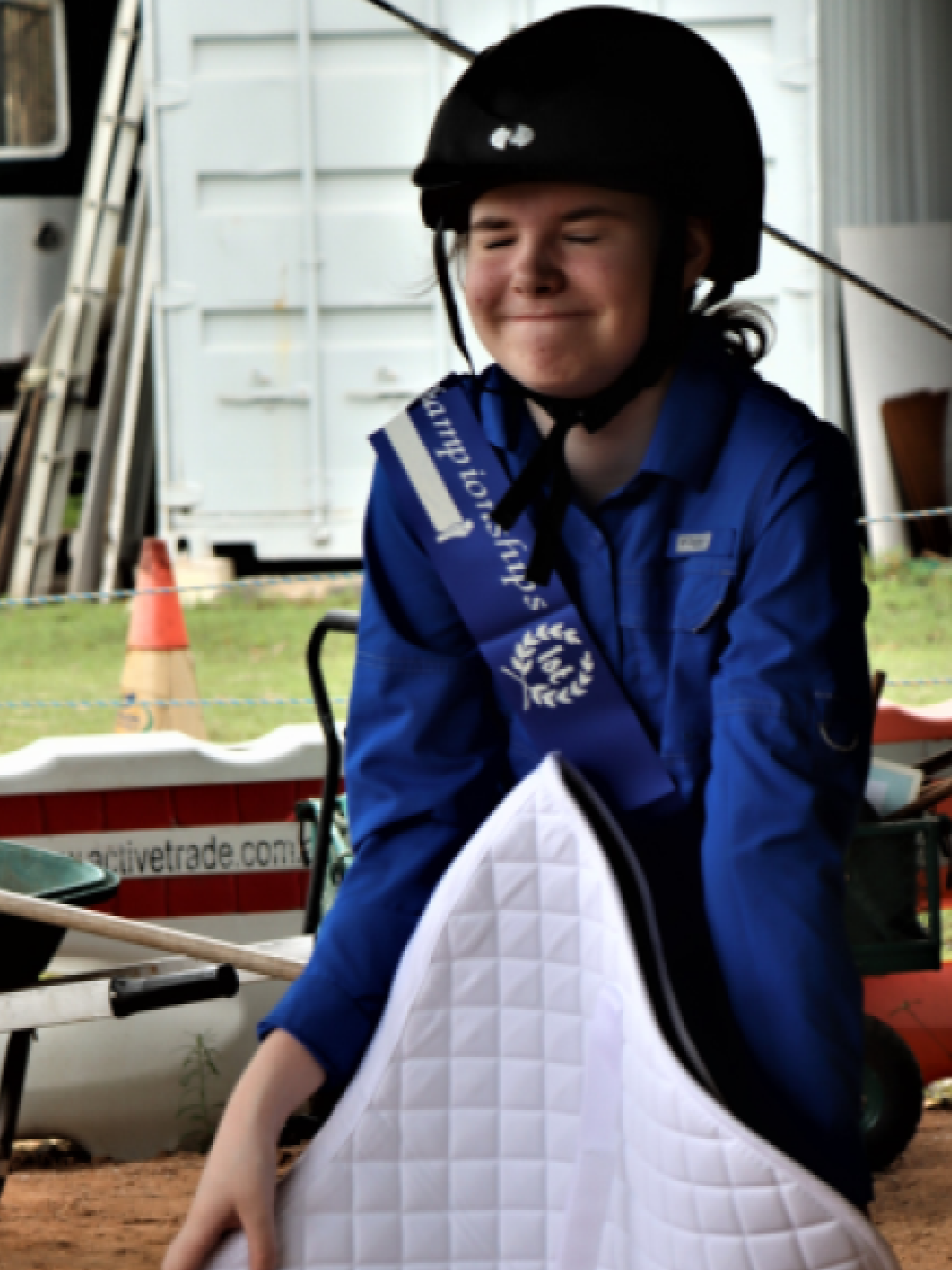 Person in blue jumpsuit and helmet holding a white saddle pad, smiling. Outdoors, near a trailer.