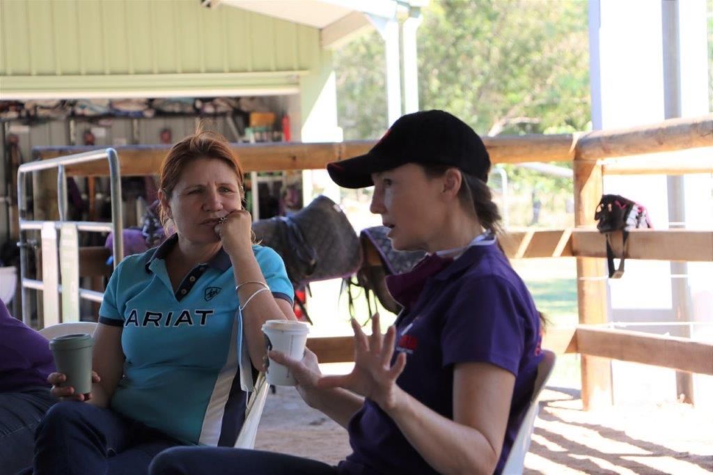 Two women in a covered outdoor area, one holding a cup, the other gesturing while talking.