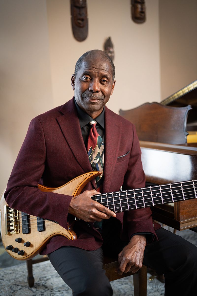 Avery Sharpe in a suit and tie is sitting on a chair holding a bass guitar .