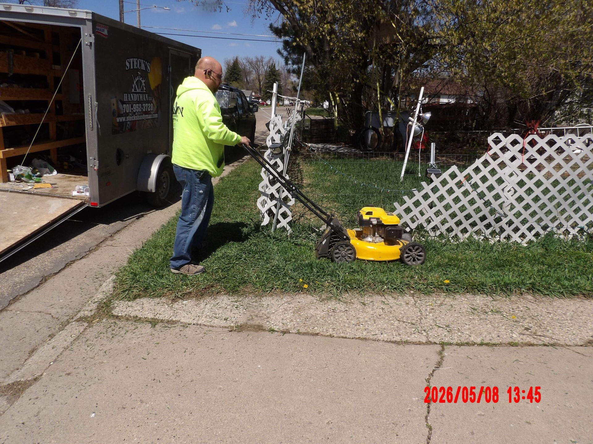Worker trimming grass beside a white fence with a yellow string trimmer and truck nearby