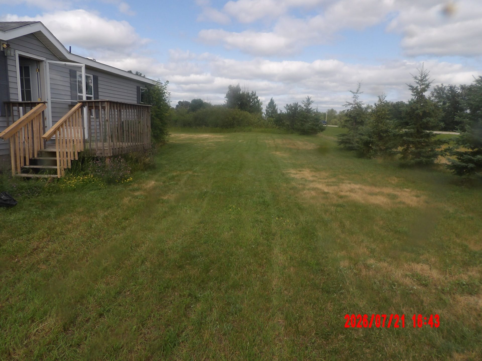 Grassy side yard beside a gray house with a wooden porch and trees under a cloudy sky