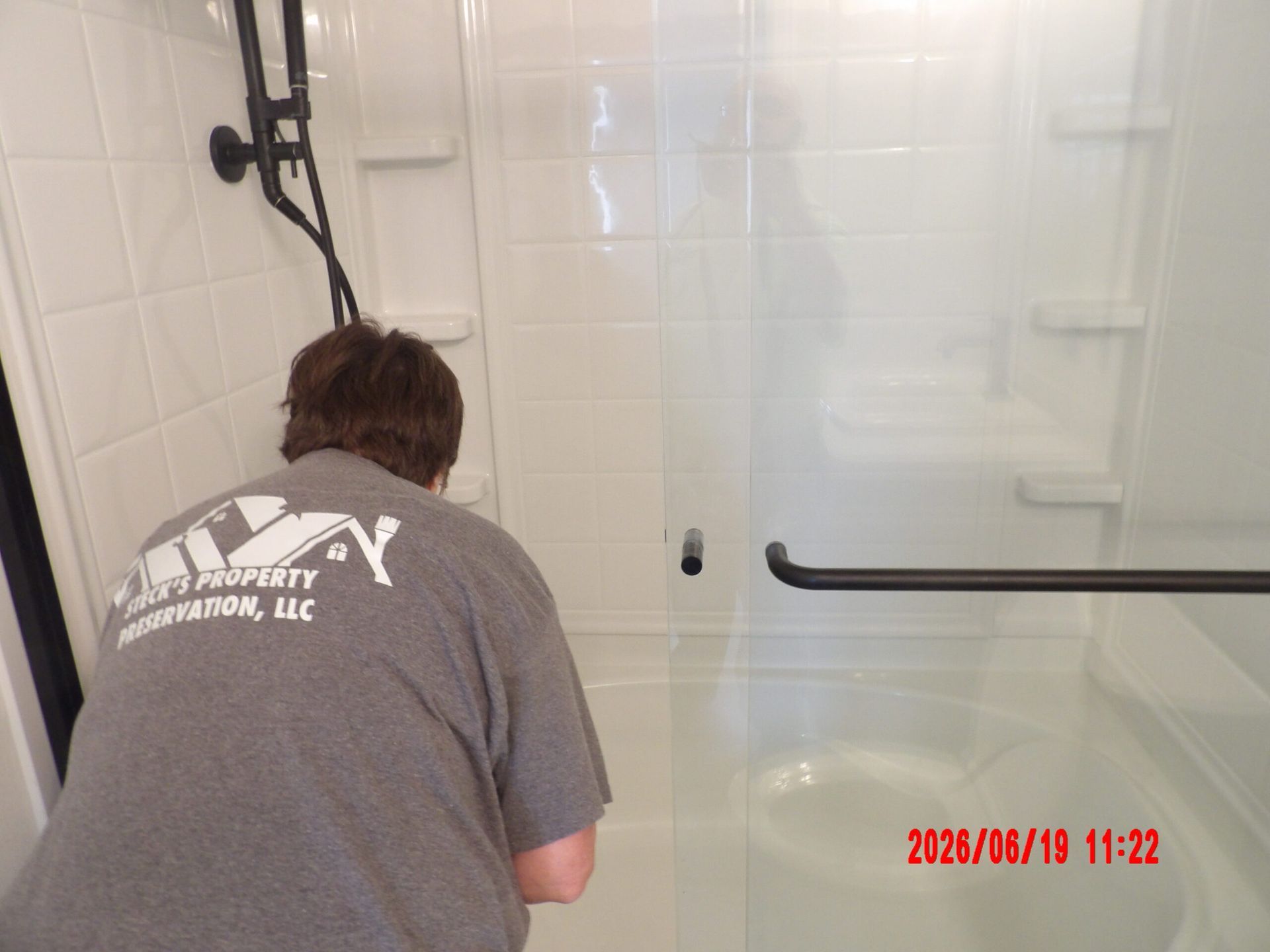 Person in gray shirt cleaning a white tiled shower stall with a black grab bar.