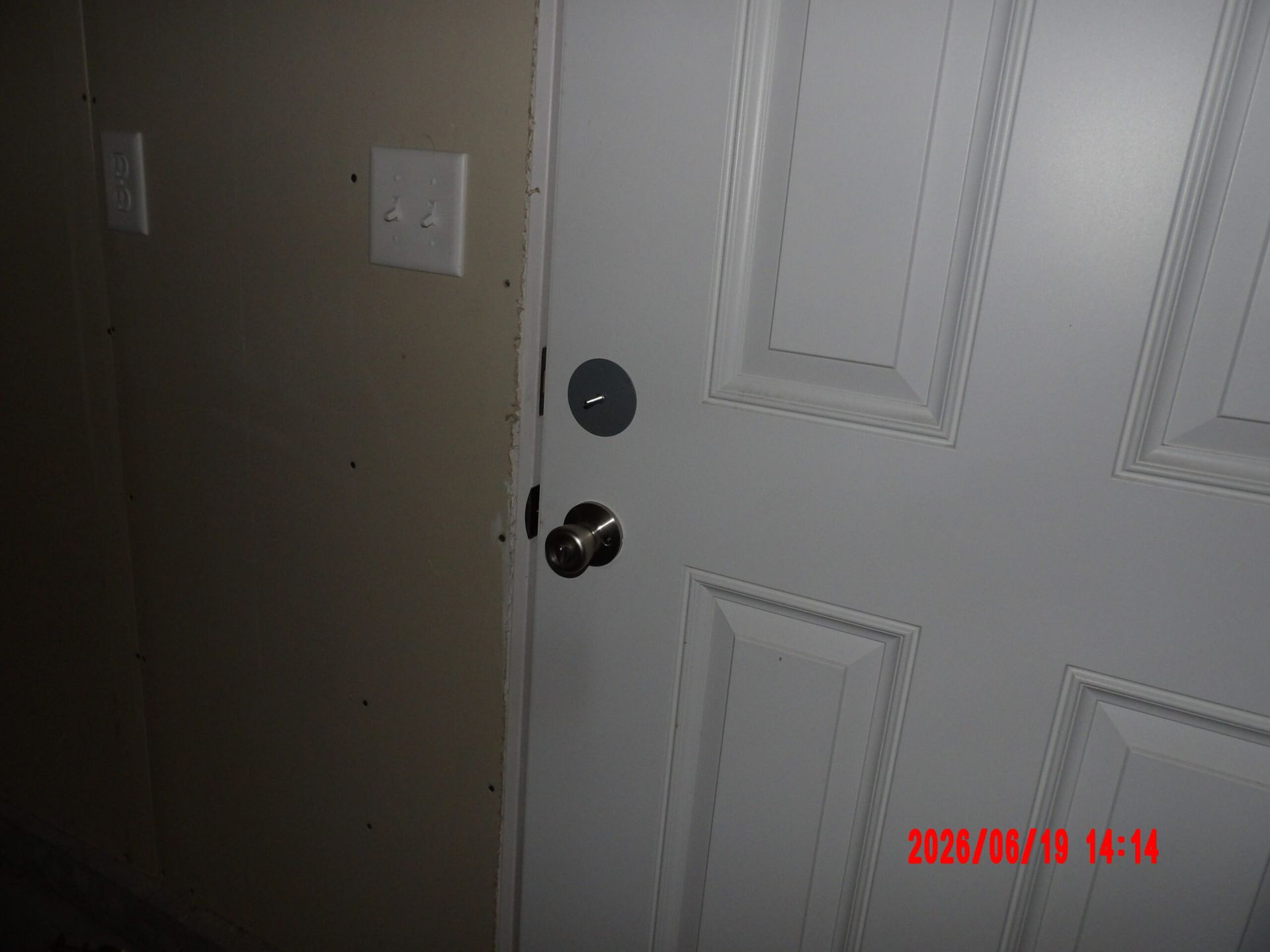 White front door with a black knob and deadbolt beside a beige wall and light switch.