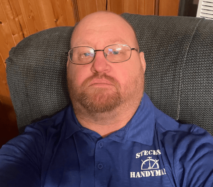 Bald man with glasses in a blue polo shirt sitting on a gray chair against wood paneling.