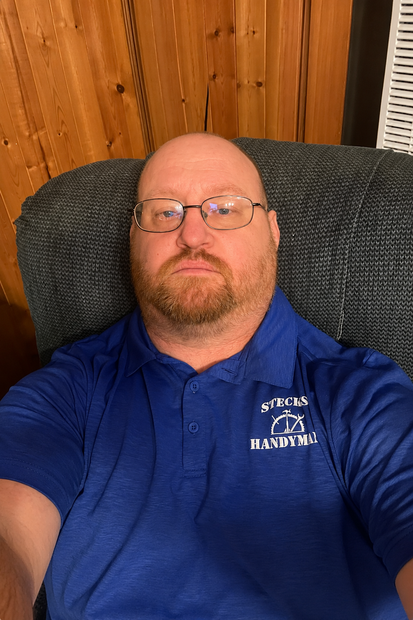 Person reclining in a gray chair, wearing a blue polo shirt and glasses, with wood-paneled wall behind them