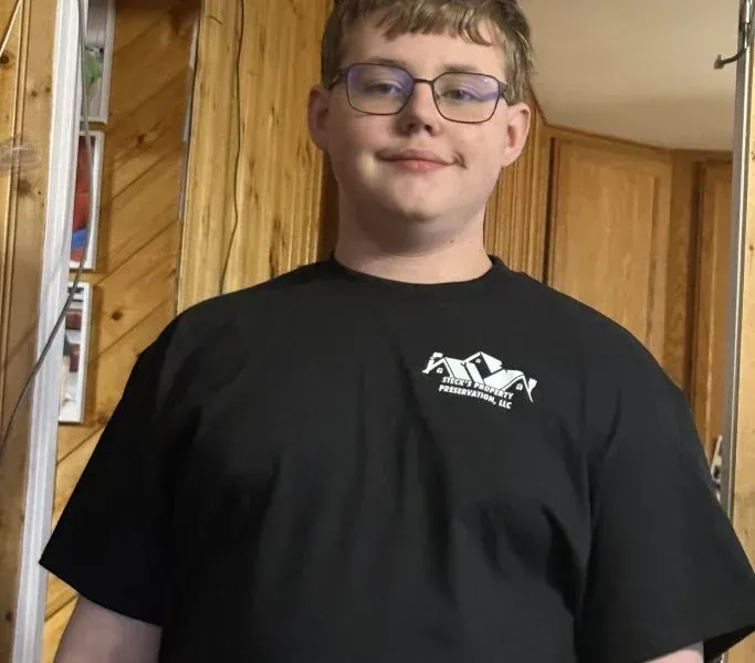 Young person in glasses wearing a black T-shirt, standing indoors against wood-paneled walls.