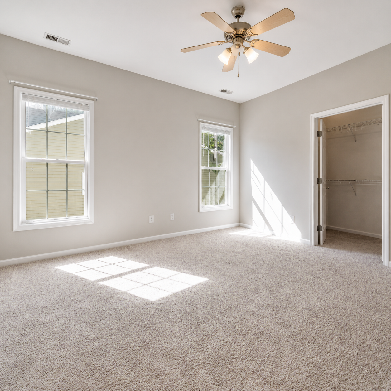 Empty beige carpeted bedroom with two windows, ceiling fan, and open closet door