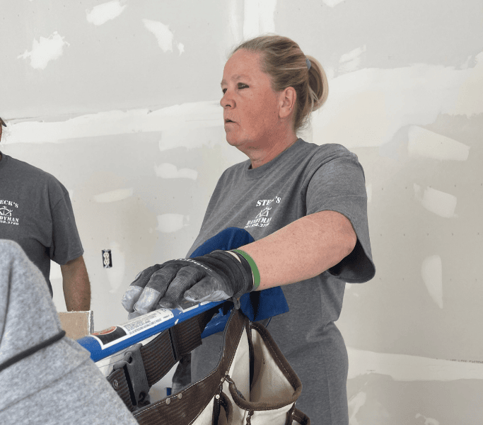 Worker in gray shirt and gloves holding a blue tool in an unfinished room with drywall patches