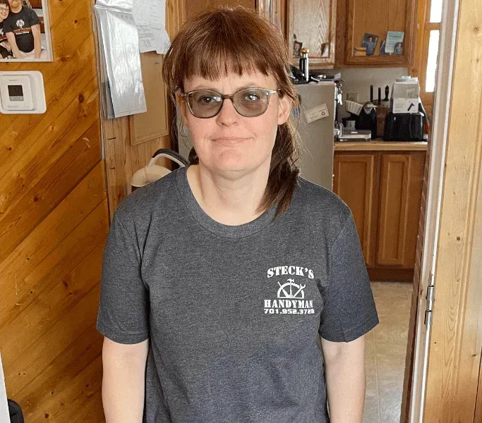 Person standing indoors in a wood-paneled kitchen, wearing glasses and a gray T-shirt.