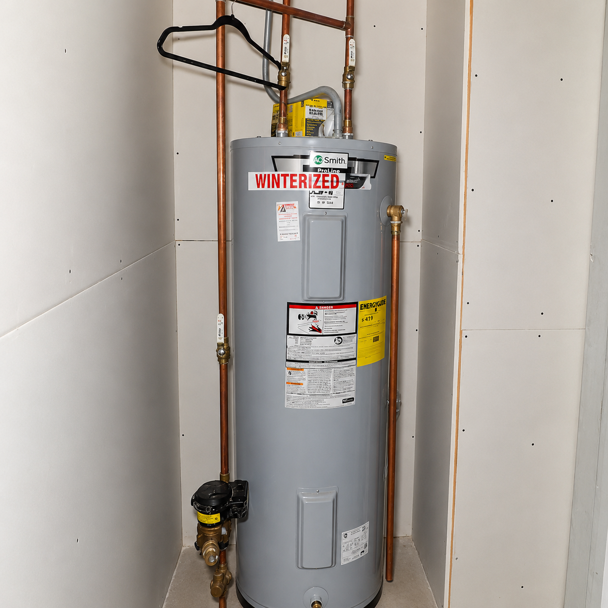 Gray water heater in a small utility closet with exposed copper pipes and warning labels.