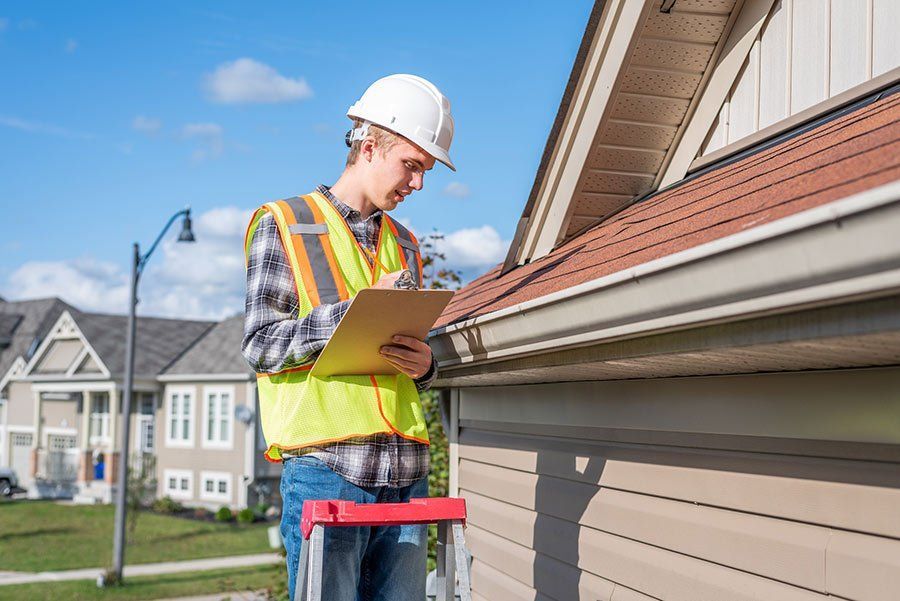 Man in safety gear on a ladder inspecting a roof, writing on a clipboard.