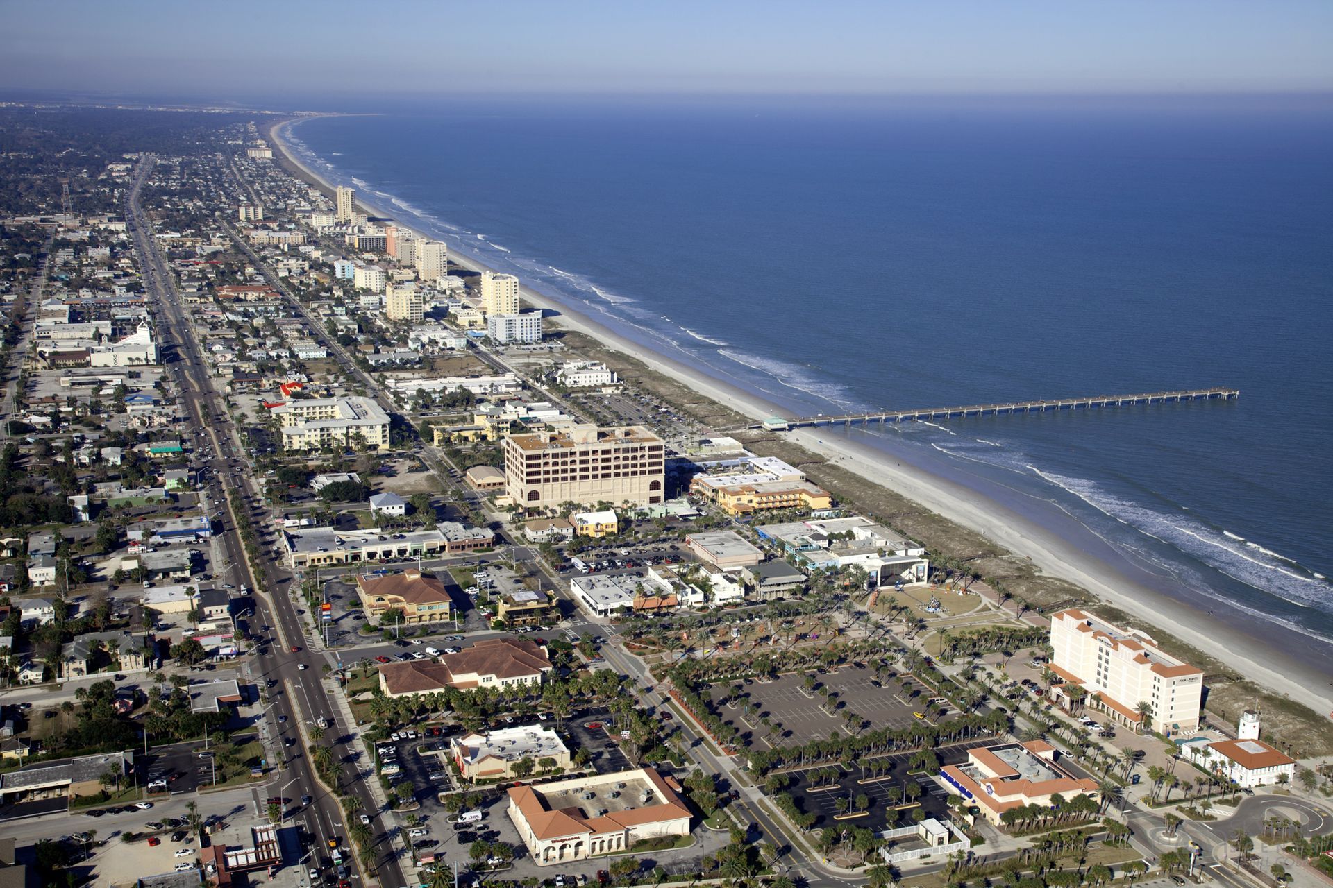 Aerial view of a coastal city with a beach and pier. Buildings line the shore, meeting the blue ocean.