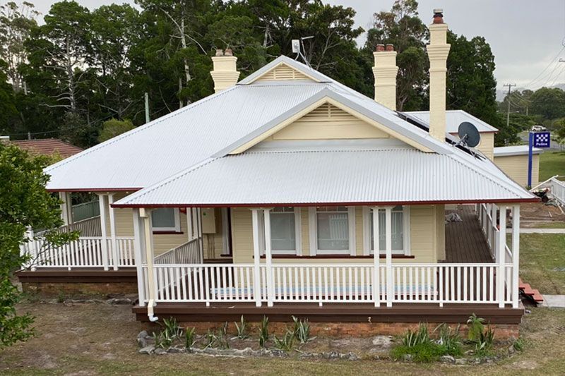 A pale yellow, single-story house with a white wraparound porch, tin roof, and tall brick chimneys.