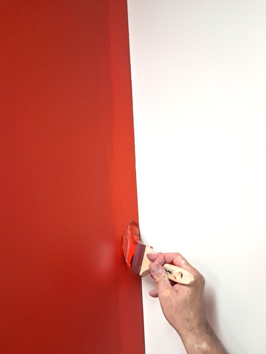 A person's hand uses a brush to paint a vertical edge on a white wall with bright red paint.