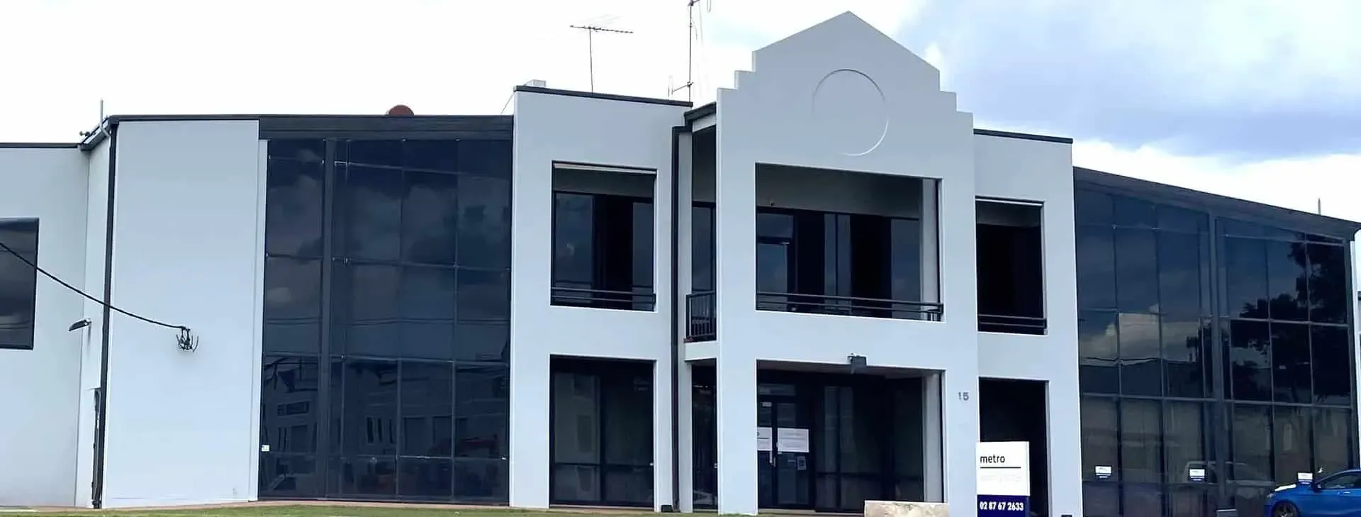 A modern, two-story commercial building featuring a white facade and dark glass paneling under a cloudy sky.