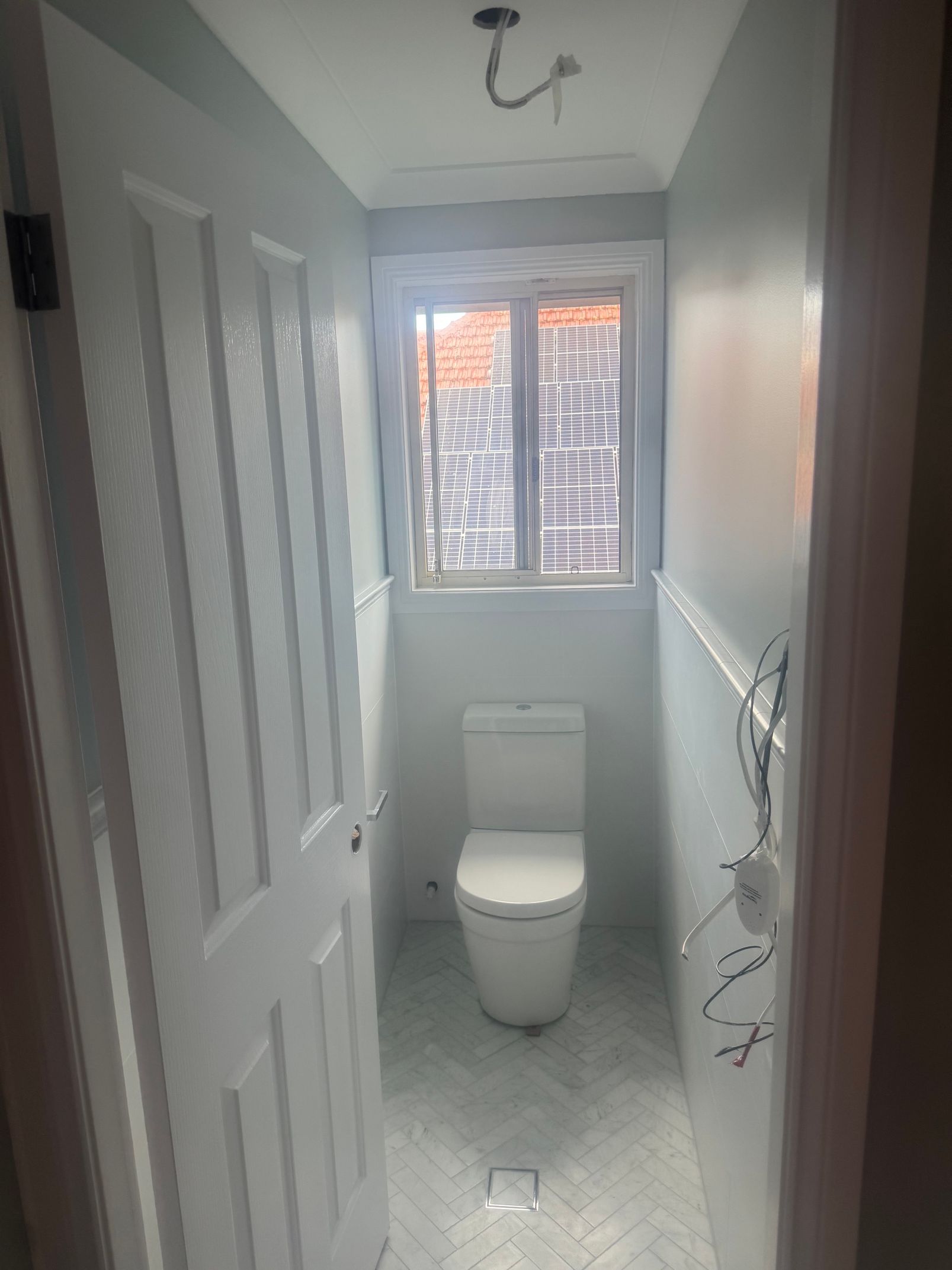 Narrow white bathroom with toilet, window, tiled floor, and unfinished light fixture overhead