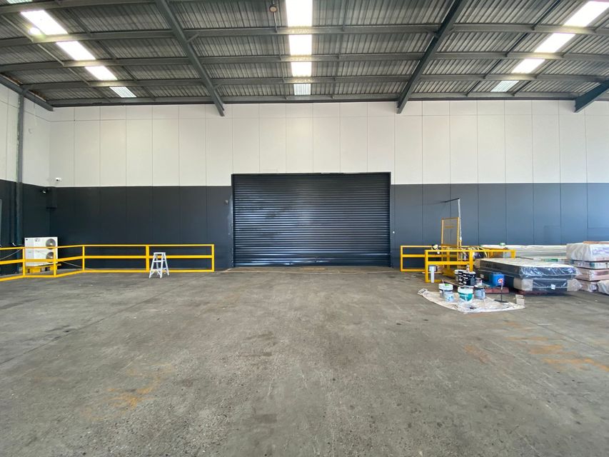 A large industrial warehouse interior with concrete floors, white and gray walls, a central roller door, and yellow rails.