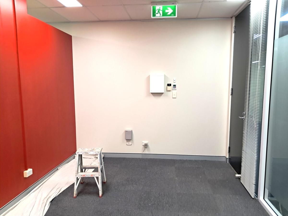 An empty room with a red wall on the left, a white wall in the center, a stepladder, and an exit sign overhead.