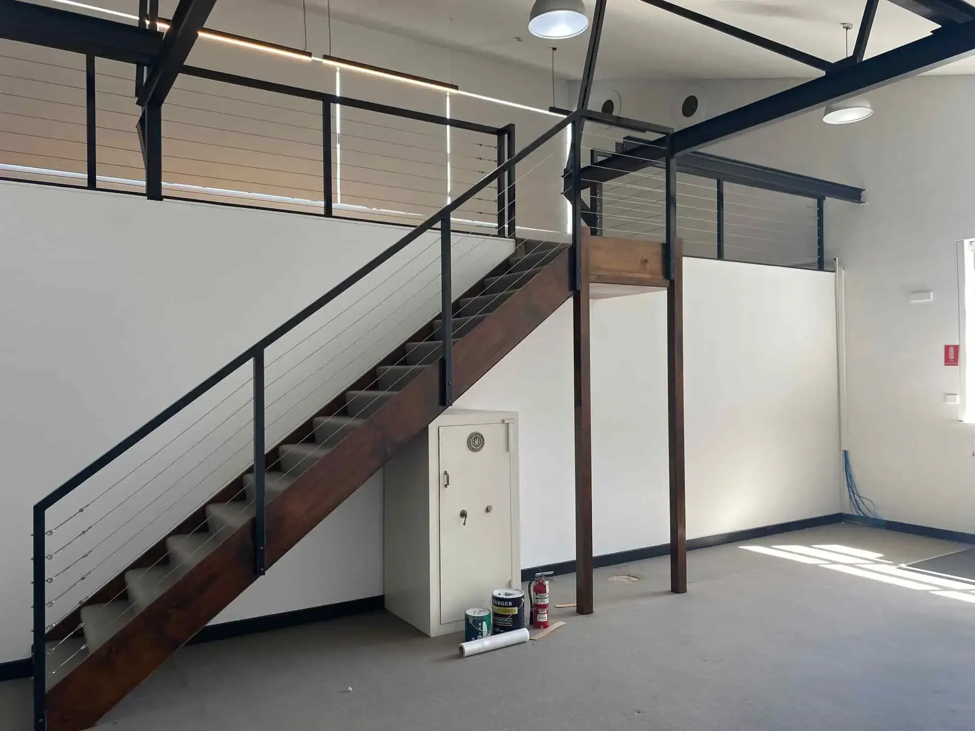 A room with a wooden staircase leading to a loft, a metal security safe underneath, and light walls with black trim.