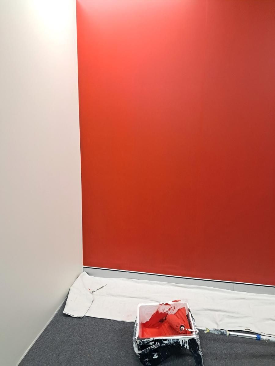 A paint tray with a roller sits on a drop cloth in front of a partially painted bright red wall in a room.