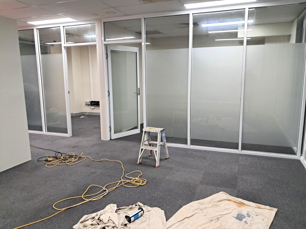 An interior office space featuring glass partitions with frosted panels, a stepladder, and tools on a grey carpeted floor.