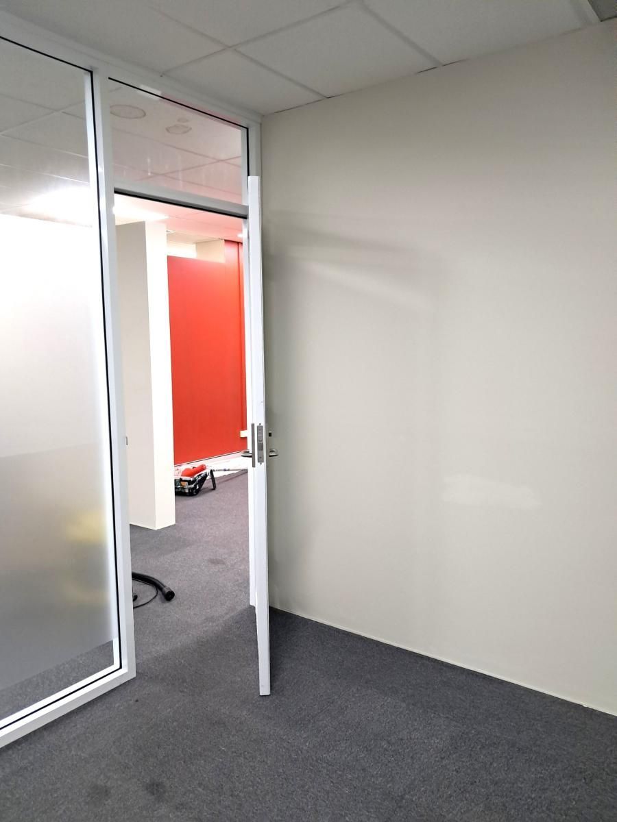 An open door in an office hallway reveals a room with a bright red wall, viewed from a grey carpeted space.