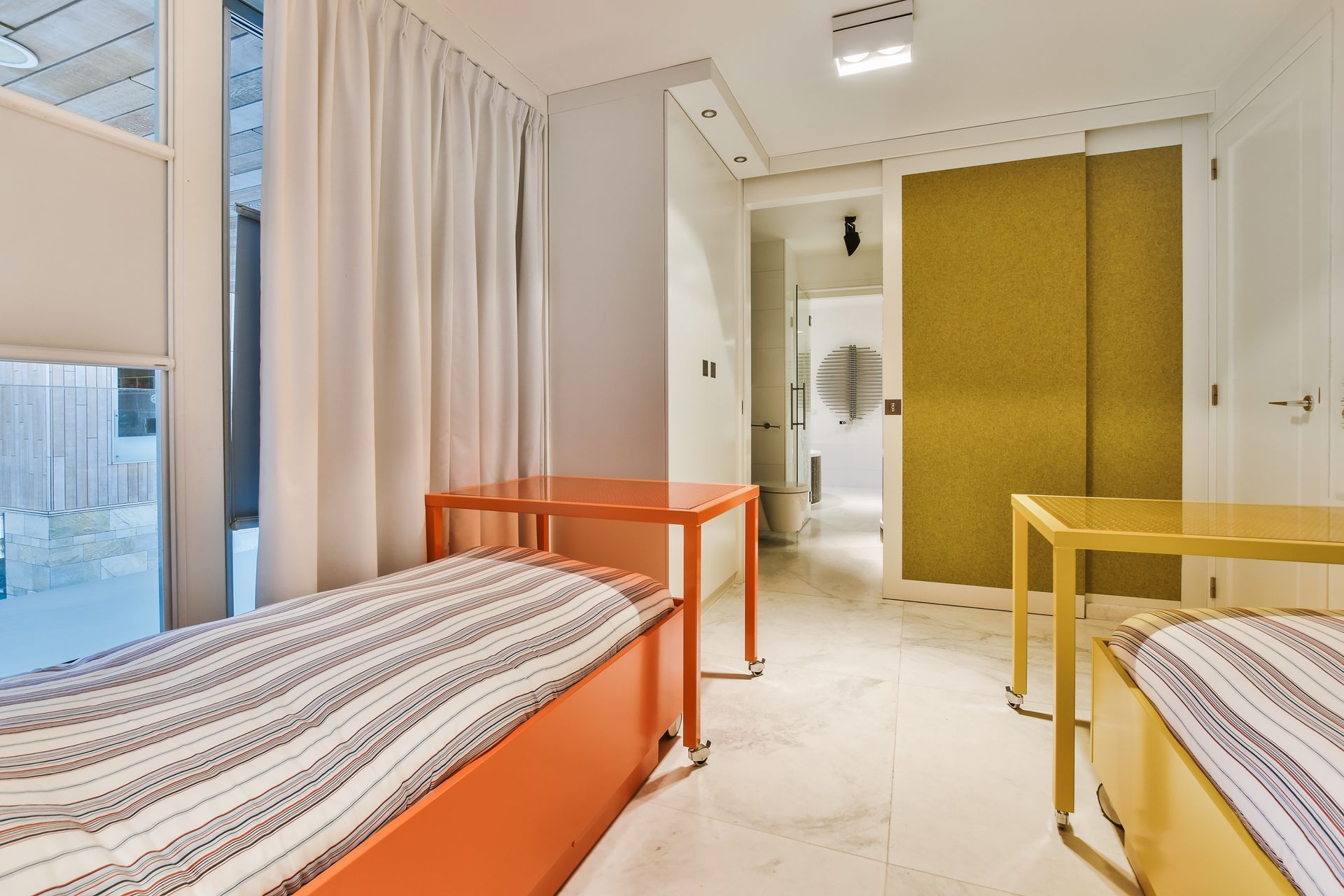 Two beds with patterned bedding next to orange and yellow tables in a room with white curtains and yellow sliding doors.