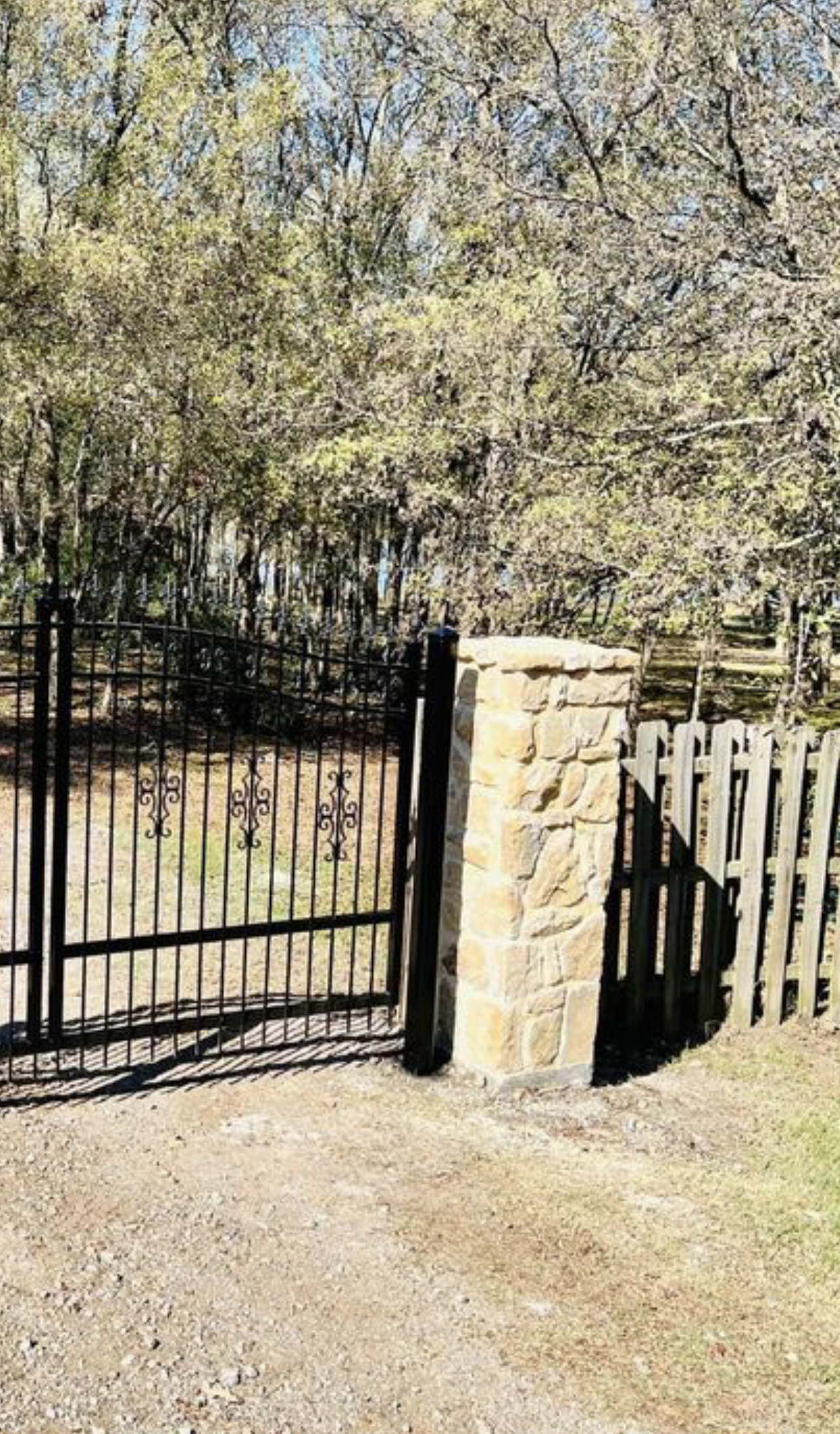A wrought iron gate is surrounded by trees and a stone pillar.
